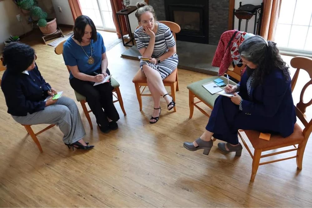 Five women sitting in a circle in conversation, notebooks in hand.